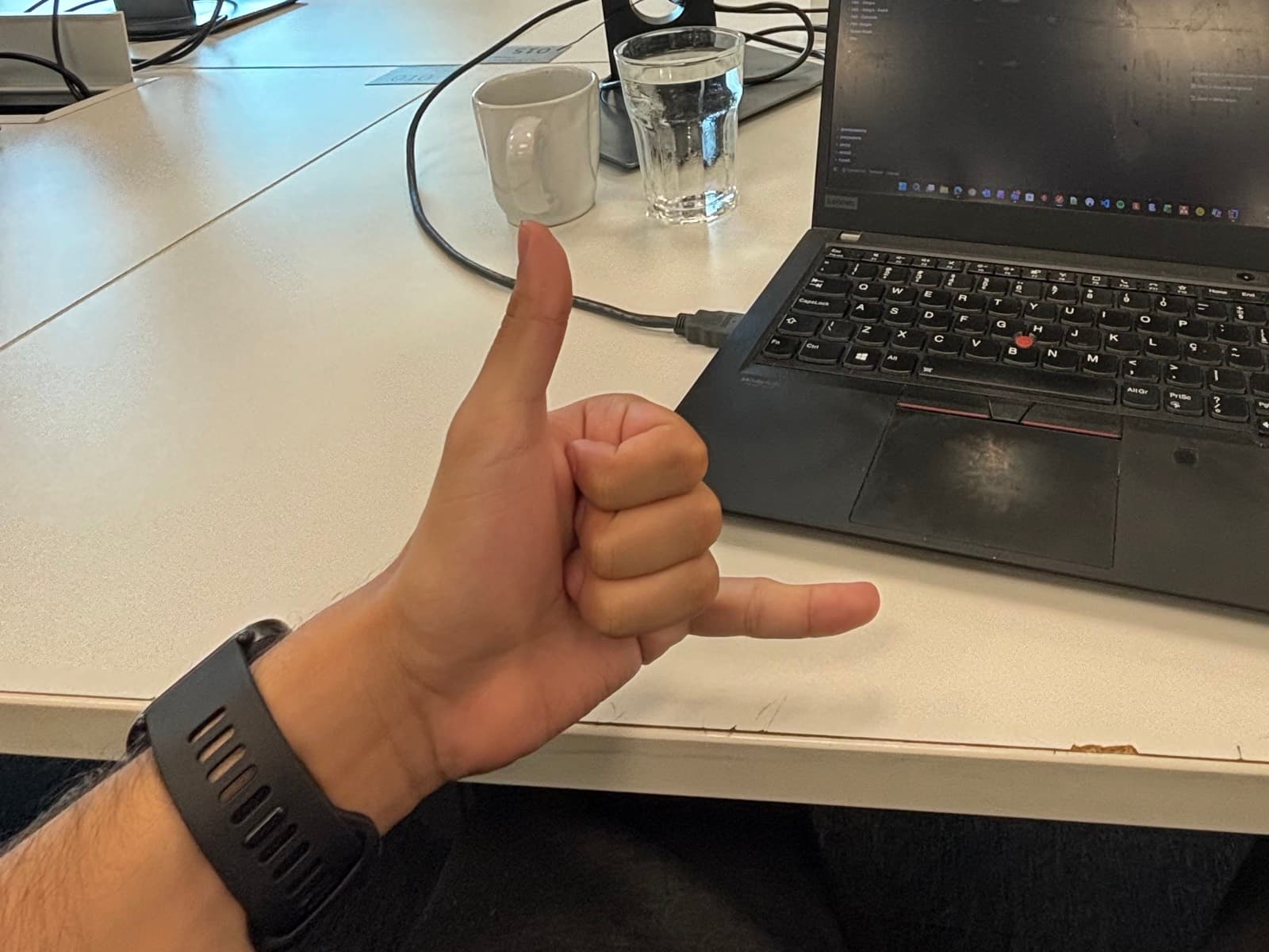 Mão fazendo o gesto de hang loose na mesa de trabalho, ao lado do notebook